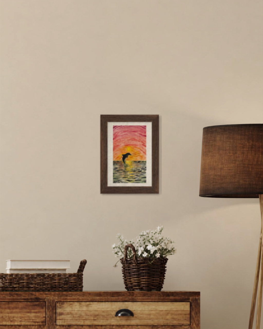 Framed mockup of the original Dolphin at Sunset watercolor painting in a brown wood frame, displayed on a wall above a console table with a lamp and fresh flowers.