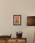 Framed mockup of the original Dolphin at Sunset watercolor painting in a brown wood frame, displayed on a wall above a console table with a lamp and fresh flowers.